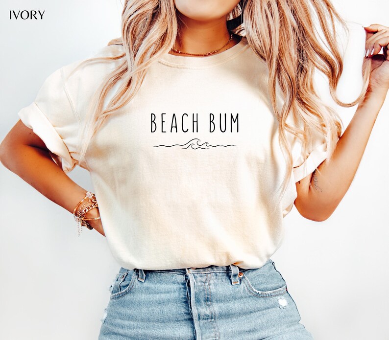 Może przedstawiać: Biały t-shirt z nadrukiem "Beach Bum" i małym wzorem fali z przodu.