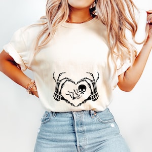 K&ouml;nnte beinhalten: Ein wei&szlig;es T-Shirt mit einem schwarzen Skelett-Hand-Design, das eine Herzform mit einem Babyskelett im Inneren bildet. Das Design ist mittig auf dem Shirt platziert.