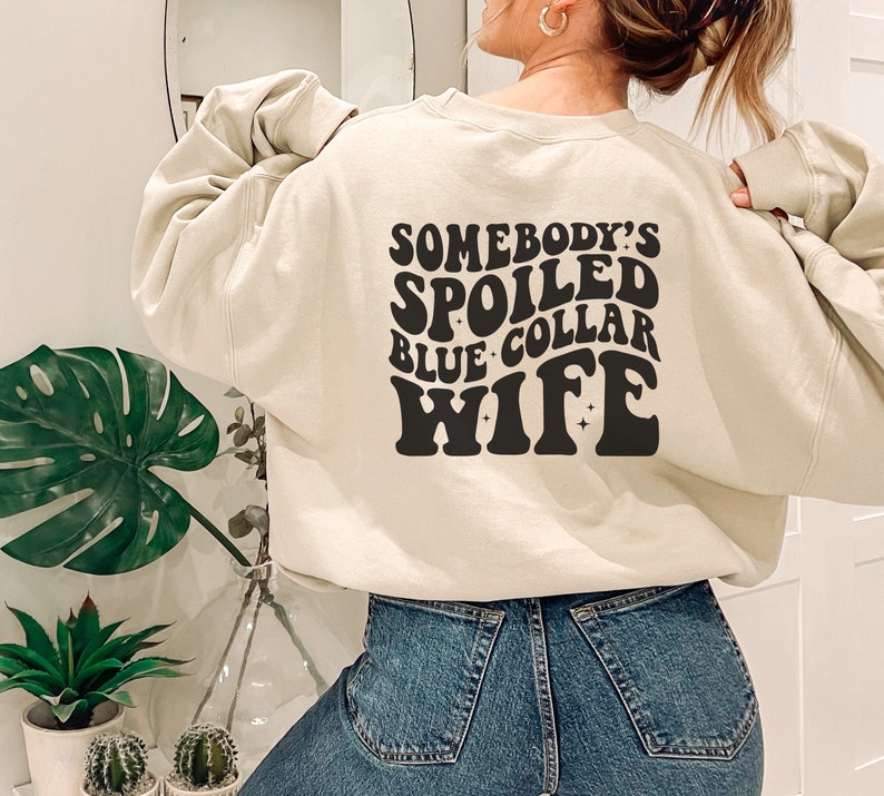 May include: A beige sweatshirt with the text "Somebody's Spoiled Blue Collar Wife" in black retro-style lettering.