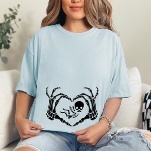 K&ouml;nnte beinhalten: Ein hellblaues T-Shirt mit einem schwarzen Skelett-Hand-Design, das eine Herzform um ein Baby-Skelett bildet. Der Text "Little Bones" ist auf das Shirt gedruckt.