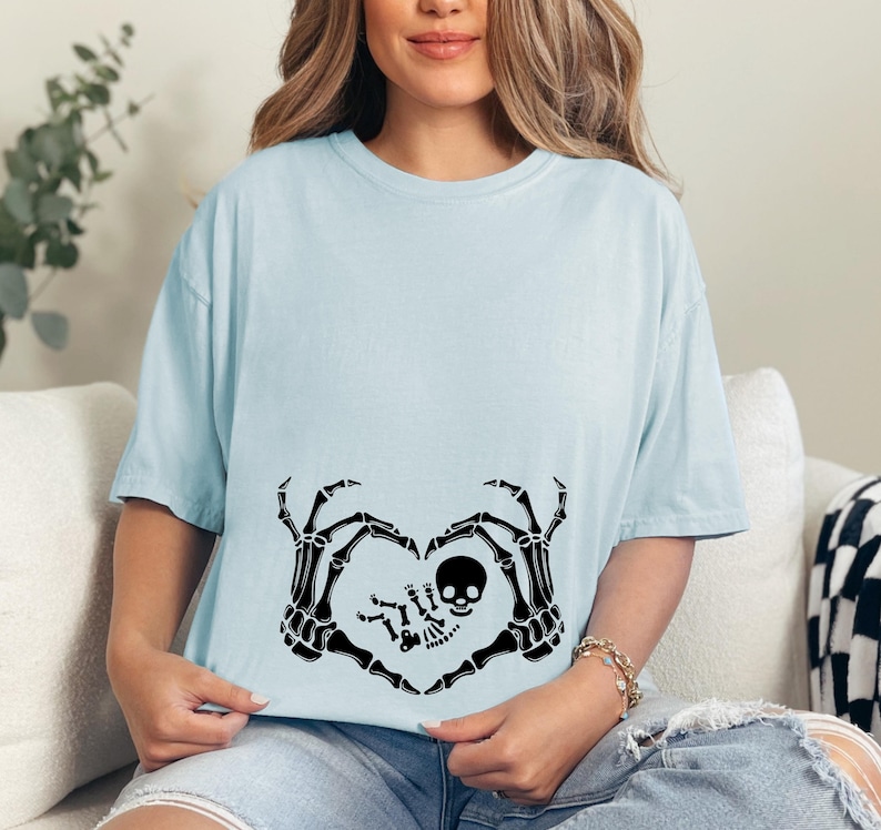 K&ouml;nnte beinhalten: Ein hellblaues T-Shirt mit einem schwarzen Skelett-Hand-Design, das eine Herzform um ein Baby-Skelett bildet. Der Text "Little Bones" ist auf das Shirt gedruckt.