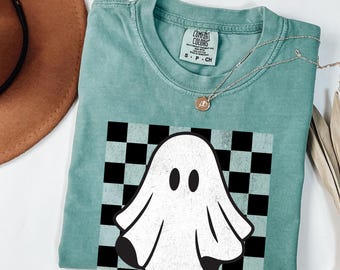 May include: Light green t-shirt featuring a white ghost graphic on a black and white chequered background. The shirt has the text "Comfort Colors" on the tag.