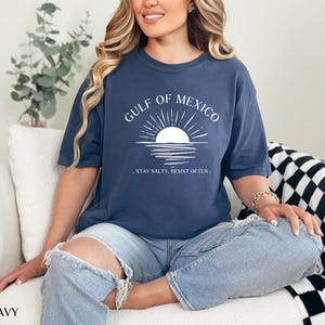 May include: A navy blue t-shirt with a white graphic of a sunset over water and the text "Gulf of Mexico. Stay Salty, Resist Often."