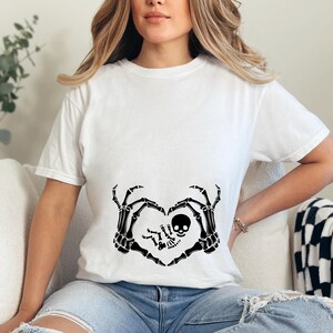 K&ouml;nnte beinhalten: Wei&szlig;es T-Shirt mit einem schwarzen Skelett-Hand-Design, das eine Herzform um ein Baby-Skelett bildet. Das Baby-Skelett tr&auml;gt eine Totenkopf-M&uuml;tze.