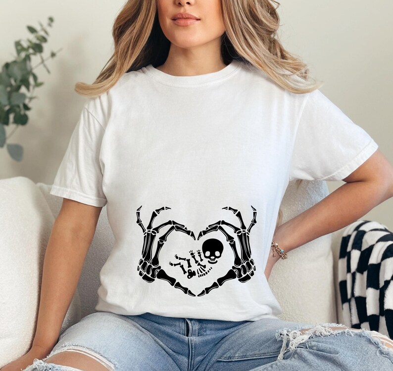 K&ouml;nnte beinhalten: Wei&szlig;es T-Shirt mit einem schwarzen Skelett-Hand-Design, das eine Herzform um ein Baby-Skelett bildet. Das Baby-Skelett tr&auml;gt eine Totenkopf-M&uuml;tze.