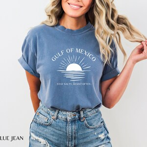 May include: A blue jean t-shirt with a white graphic of a sunset over the Gulf of Mexico. The text "Stay Salty, Resist Often" is printed below the graphic.