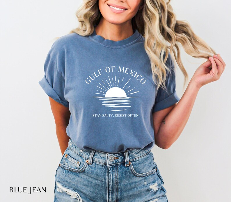May include: A blue jean t-shirt with a white graphic of a sunset over the Gulf of Mexico. The text "Stay Salty, Resist Often" is printed below the graphic.