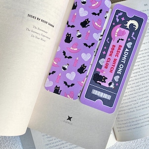 May include: Two decorative bookmarks with a witchy theme rest on the pages of a book. One bookmark is purple with black cats, witch hats, and bats. The other is a ticket-shaped bookmark with the text "BASIC WITCH BOOK CLUB".