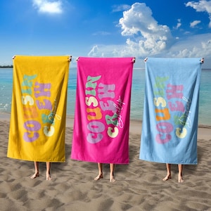 May include: Three beach towels in yellow, pink, and blue, each with the text "COUSIN CREW" in a retro font. The towels are held up on a sandy beach with the ocean and sky in the background.