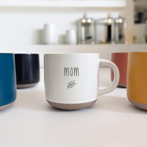 May include: A collection of ceramic mugs in various colors, including blue, black, white, coral, and yellow. The white mug features the word "mom" and a leaf design. The mugs have a speckled, textured appearance and a neutral-colored base.