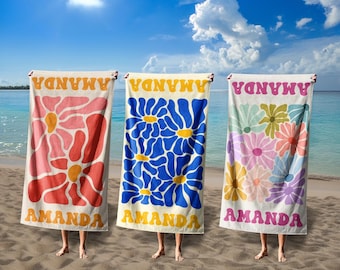 May include: Three beach towels with floral designs and the name "AMANDA" printed on them. One towel has a red flower, another has blue flowers, and the third has a mix of pastel flowers. The towels are held up on a sandy beach with the ocean in the background.