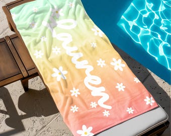 May include: A beach towel with a gradient of colors, from light green to yellow to orange, with the name "Lauren" in white script. White daisy flowers are scattered across the towel. The towel is on a wooden lounge chair.