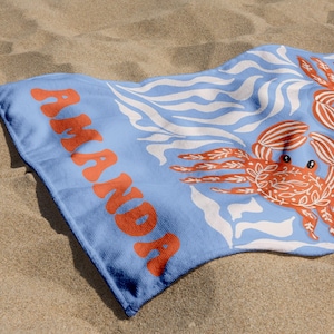 Puede incluir: Una toalla de playa azul claro con el nombre "AMANDA" en letras naranjas. La toalla presenta un patr&oacute;n de l&iacute;neas onduladas blancas y dos ilustraciones de cangrejos naranjas. La toalla est&aacute; sobre una superficie arenosa.
