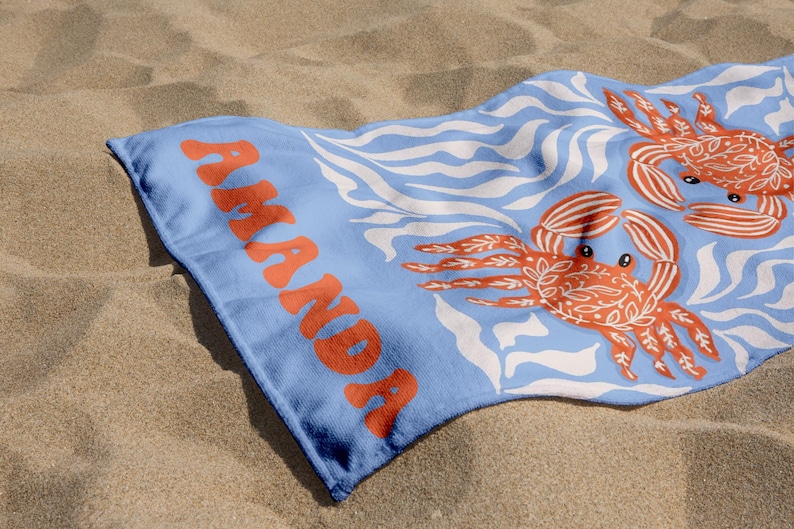 Puede incluir: Una toalla de playa azul claro con el nombre "AMANDA" en letras naranjas. La toalla presenta un patr&oacute;n de l&iacute;neas onduladas blancas y dos ilustraciones de cangrejos naranjas. La toalla est&aacute; sobre una superficie arenosa.