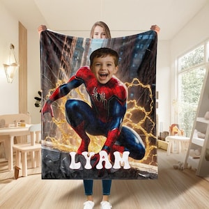 May include: A personalized blanket featuring a child's face superimposed on a Spider-Man costume. The blanket has a cityscape background with the name "LYAM" in white block letters at the bottom. The child is smiling.