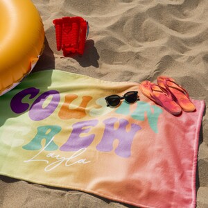 May include: A beach scene with a colorful towel, sunglasses, and flip-flops. The towel has the words "COAST CREW Layla" in a retro font. An inflatable yellow ring and a red bucket are also present, all on a sandy surface.