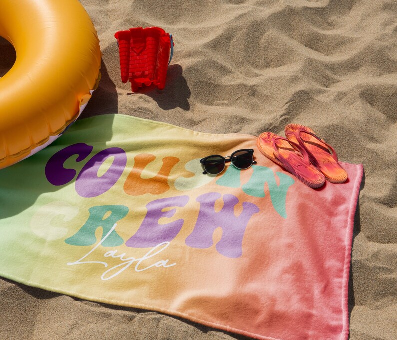 May include: A beach scene with a colorful towel, sunglasses, and flip-flops. The towel has the words "COAST CREW Layla" in a retro font. An inflatable yellow ring and a red bucket are also present, all on a sandy surface.