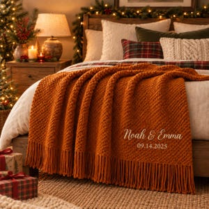May include: A burnt orange knitted throw blanket with fringe trim, featuring the embroidered text "Noah & Emma" and the date "14.09.2025" in a cursive font. The blanket has a textured diagonal stitch pattern and is draped over a bed.