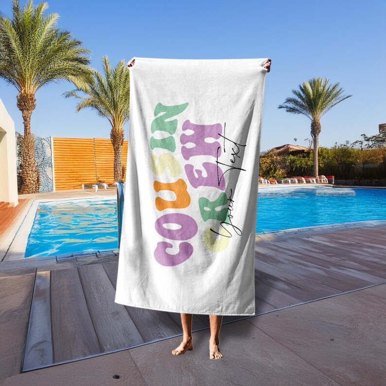 May include: White beach towel with the words "COUSIN CREW" in colorful, retro-style lettering. The towel is held up in front of a pool with palm trees in the background. The words "Your Text" are also on the towel.