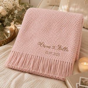 May include: A soft, light pink knitted throw blanket featuring a textured popcorn stitch pattern and long fringe along the bottom edge. The blanket is embroidered with the names Owen and Stella separated by a heart, and the date 12.07.2025.