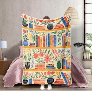May include: A cream-coloured throw blanket with a colourful bookshelf design. The design features stacks of books, potted plants, and flowers in shades of red, blue, orange, and green. The blanket is held up by a person.