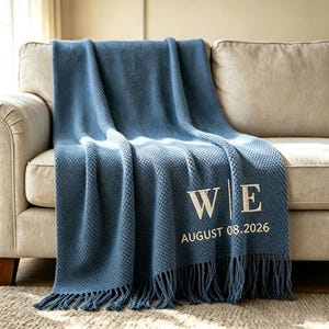 Pode incluir: Uma manta de malha azul com um padrão de textura de waffle e franjas. A manta é bordada com as letras W e E separadas por uma linha vertical, com o texto AUGUST 08.2026 posicionado diretamente abaixo das letras bordadas na manta.