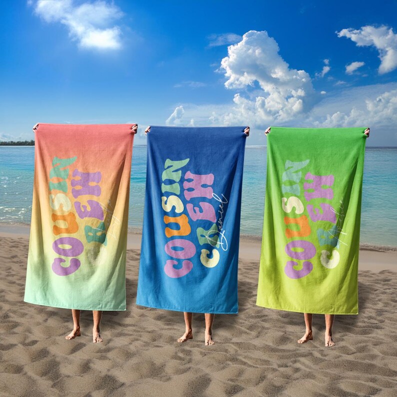 May include: Three beach towels with the text "Cousin Crew" in a retro font. The towels are held up on a sandy beach with the ocean and sky in the background. The towels are in shades of orange, blue, and green.