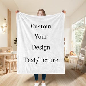 Op de afbeelding: Een witte deken met de tekst "Custom Your Design Text/Picture" in het zwart. De deken wordt in een lichte kamer omhoog gehouden. Het is een aanpasbaar product.