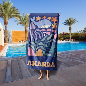 Puede incluir: Toalla de playa azul marino con un dise&ntilde;o oce&aacute;nico caprichoso que presenta una ballena, medusas, estrellas de mar y peces en tonos azules, rosas y naranjas. El nombre "AMANDA" est&aacute; impreso en amarillo.