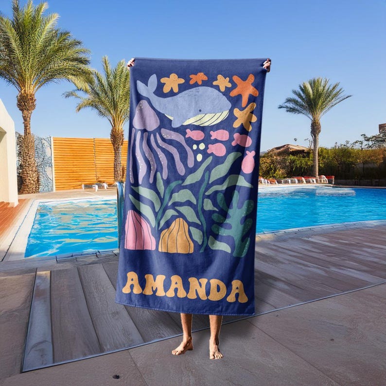 Puede incluir: Toalla de playa azul marino con un dise&ntilde;o oce&aacute;nico caprichoso que presenta una ballena, medusas, estrellas de mar y peces en tonos azules, rosas y naranjas. El nombre "AMANDA" est&aacute; impreso en amarillo.