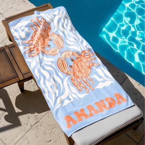 Puede incluir: Una toalla de playa azul claro con dos ilustraciones de cangrejos naranjas y el nombre "AMANDA" en letras naranjas. La toalla presenta un patr&oacute;n de olas en blanco y est&aacute; colocada sobre una tumbona de madera junto a una piscina.