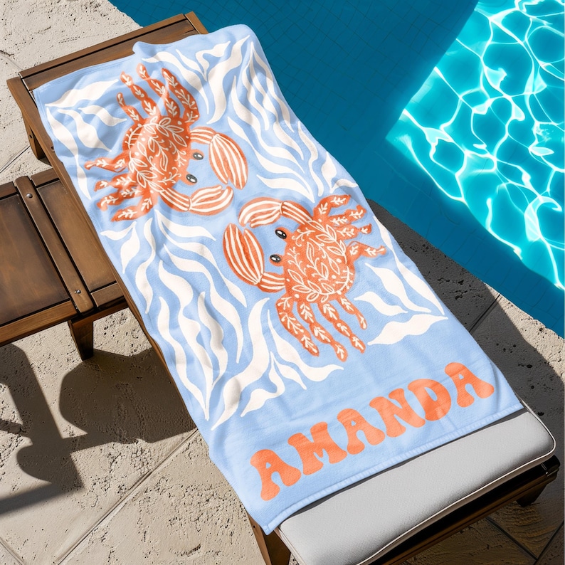Puede incluir: Una toalla de playa azul claro con dos ilustraciones de cangrejos naranjas y el nombre "AMANDA" en letras naranjas. La toalla presenta un patr&oacute;n de olas en blanco y est&aacute; colocada sobre una tumbona de madera junto a una piscina.