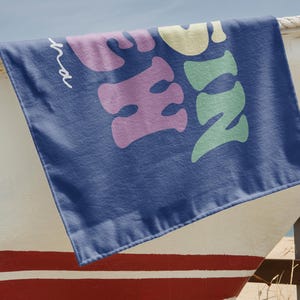 May include: A navy blue beach towel with the word "Sunshine" in large, colorful, bubble letters. The towel is draped over a white and red painted boat. The text is in shades of pink, yellow, and green.