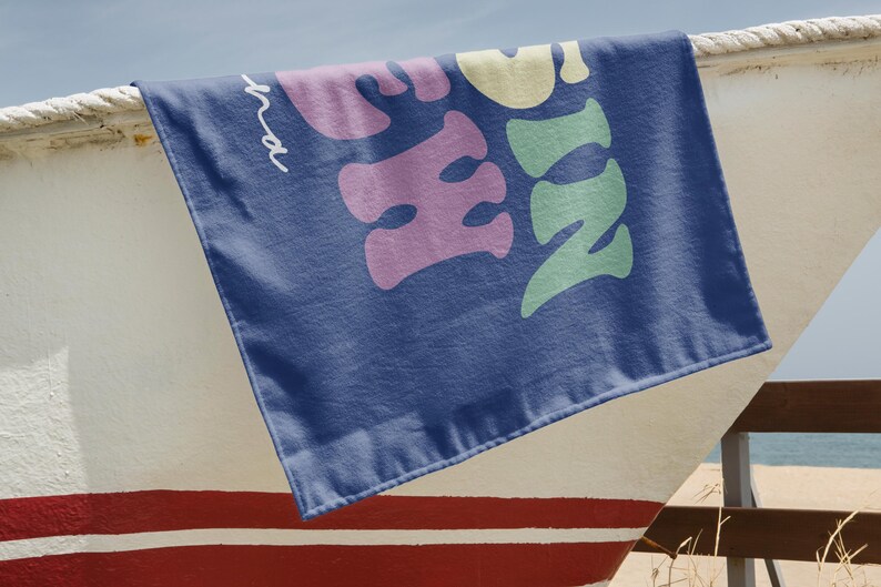 May include: A navy blue beach towel with the word "Sunshine" in large, colorful, bubble letters. The towel is draped over a white and red painted boat. The text is in shades of pink, yellow, and green.
