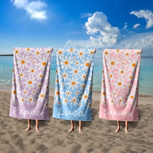 May include: Three beach towels with a daisy print. The towels are pink, blue, and purple. Each towel has a name written on it: Pepper, Luca, and Sakura.
