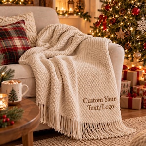 May include: A cream-coloured knit throw blanket with a textured pattern and long fringe trim. The blanket features the embroidered text "Custom Your Text/Logo" in a brown font, draped over a sofa next to a plaid pillow and a mug.
