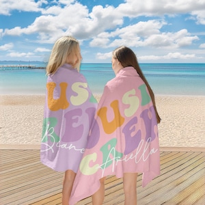 May include: Two beach towels with the text "US" in orange, "S" in yellow, "E" in green, "Y" in purple, and names in white script. One towel is lavender, the other is pink. The towels are on a wooden deck with a beach and ocean background.