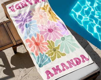 May include: A white beach towel with a colorful floral design and the name "AMANDA" in pink lettering. The towel features large, stylized flowers in shades of pink, purple, orange, yellow, and blue. The towel is draped over a wooden lounge chair.
