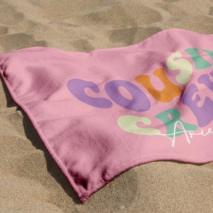 May include: A pink beach towel with the words "Cousin Crew" in colorful, retro-style lettering. The name "Ariella" is written in cursive below the text. The towel is lying on a sandy surface.