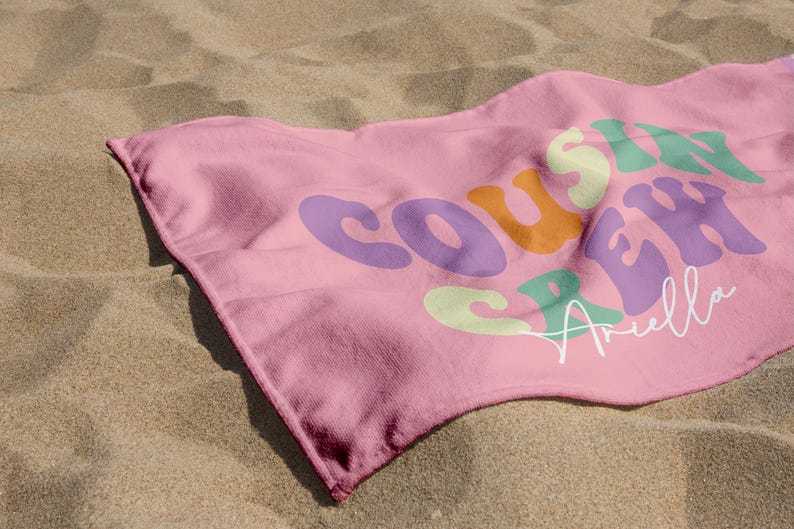 May include: A pink beach towel with the words "Cousin Crew" in colorful, retro-style lettering. The name "Ariella" is written in cursive below the text. The towel is lying on a sandy surface.