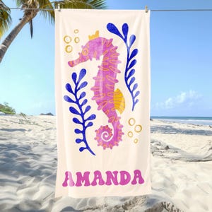 Puede incluir: Una toalla de playa crema con un dise&ntilde;o de caballito de mar en tonos rosa, amarillo y azul. El caballito de mar est&aacute; rodeado de algas azules y burbujas doradas. El nombre "AMANDA" est&aacute; impreso en rosa en la parte inferior.