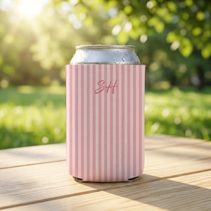 Op de afbeelding: Een roze en witte gestreepte blikjeskoeler met de initialen "SH" in een cursief lettertype. De koeler staat op een houten oppervlak, met een blikje drank erin. De achtergrond is een wazig beeld van groen gras en bomen.