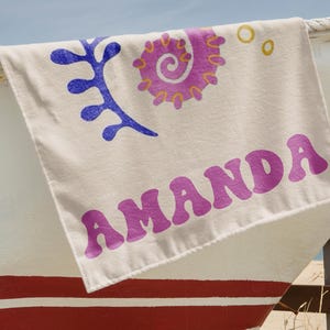 Puede incluir: Una toalla de playa color crema con el nombre "AMANDA" en letras moradas estilizadas. La toalla presenta dise&ntilde;os abstractos azules y morados. La toalla est&aacute; sobre un barco blanco y rojo.