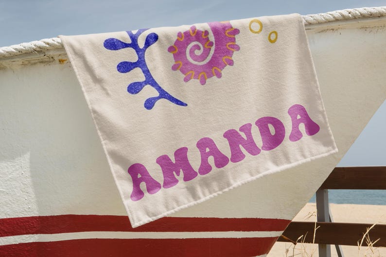 Puede incluir: Una toalla de playa color crema con el nombre "AMANDA" en letras moradas estilizadas. La toalla presenta dise&ntilde;os abstractos azules y morados. La toalla est&aacute; sobre un barco blanco y rojo.