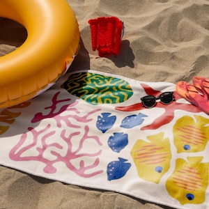 Puede incluir: Escena de playa con un flotador inflable amarillo, un cubo de arena rojo, una toalla de playa estampada, gafas de sol y chanclas. La toalla presenta ilustraciones coloridas con tem&aacute;tica marina, ideal para un d&iacute;a en la playa.