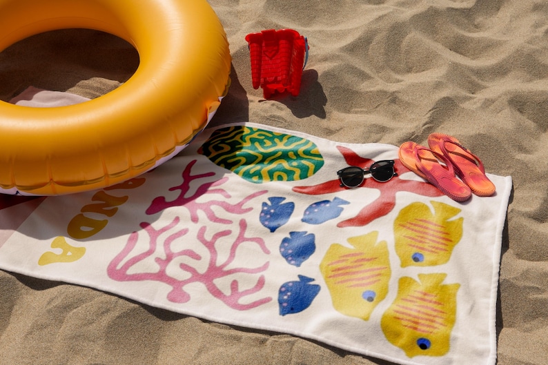 Puede incluir: Escena de playa con un flotador inflable amarillo, un cubo de arena rojo, una toalla de playa estampada, gafas de sol y chanclas. La toalla presenta ilustraciones coloridas con tem&aacute;tica marina, ideal para un d&iacute;a en la playa.