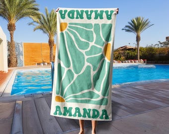 May include: A white beach towel with a teal floral design and the name "AMANDA" in green. The towel is held up in front of a pool with palm trees in the background.