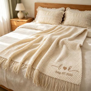 Può includere: Una coperta in maglia color crema con un motivo a bolle testurizzato e bordi con lunghe frange. La coperta presenta un testo ricamato in marrone che recita L cuore K e la data Aug. 07.2026, centrato sulla parte inferiore del tessuto.