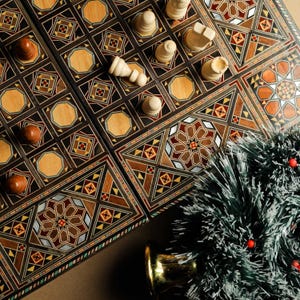 May include: A wooden backgammon board with intricate inlaid designs in shades of brown, red, and gold. White game pieces are arranged on the board. A small artificial Christmas tree with red berries sits in the bottom right corner of the image.