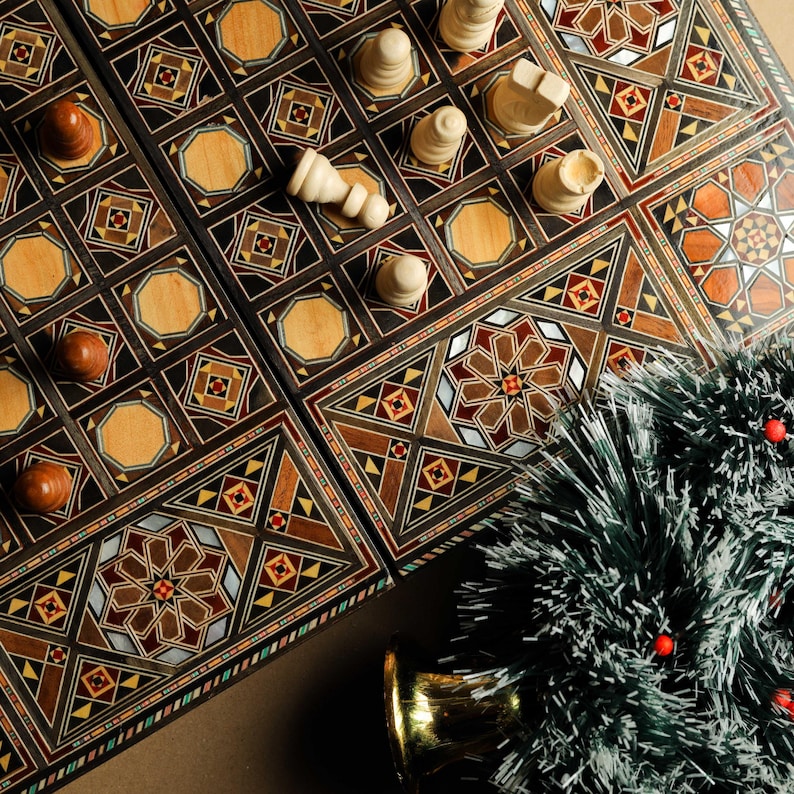 May include: A wooden backgammon board with intricate inlaid designs in shades of brown, red, and gold. White game pieces are arranged on the board. A small artificial Christmas tree with red berries sits in the bottom right corner of the image.